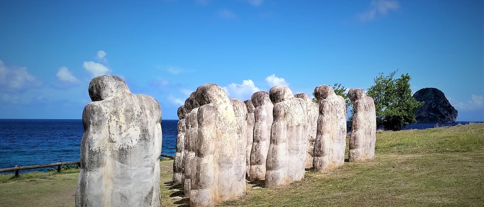 Les statues blanches du Mémorial Cap 110 alignées face à la mer à l’Anse Caffard, au Diamant, avec le Rocher du Diamant en arrière-plan